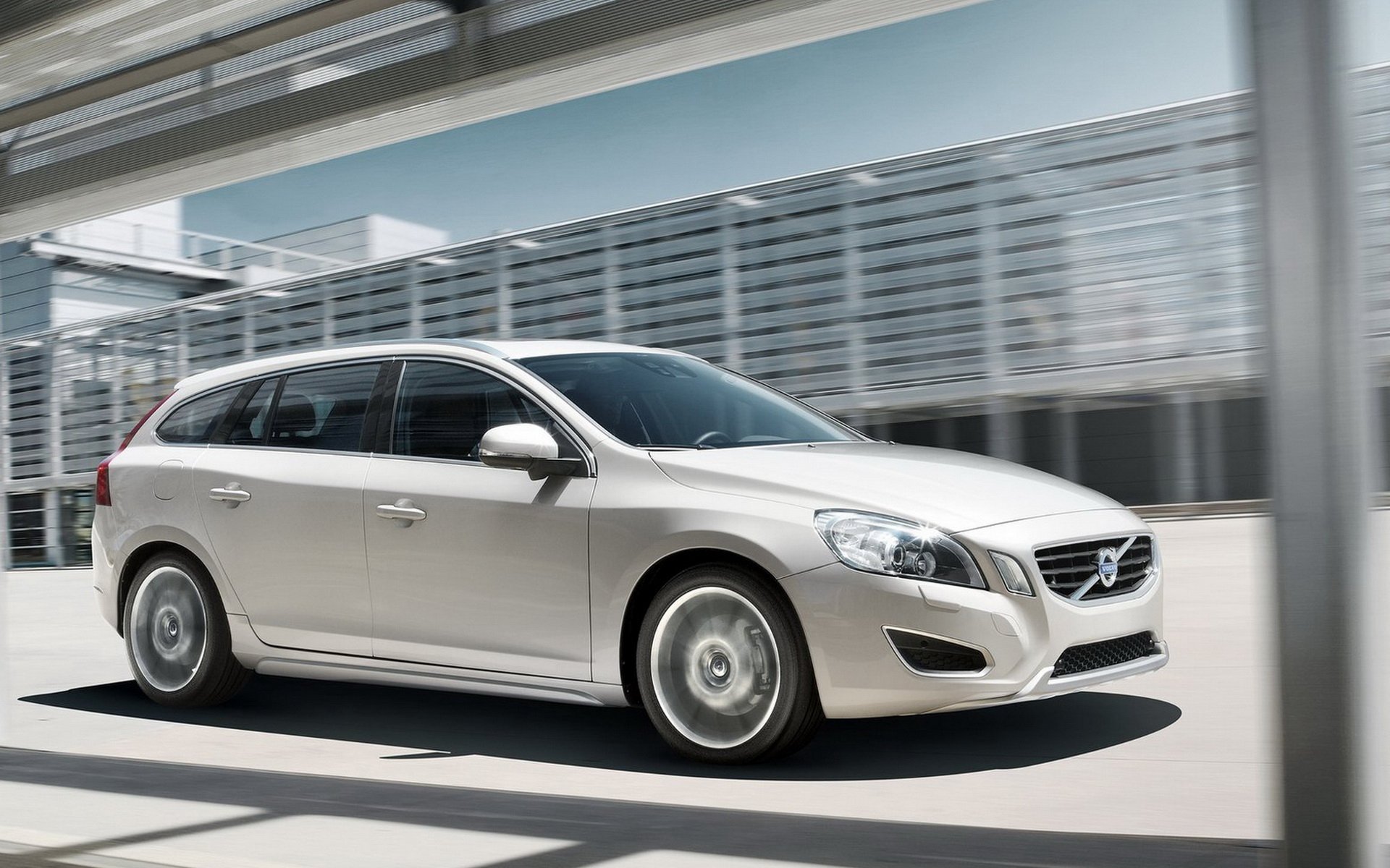 Vehicle: silver Volvo V60 wagon in motion by a modern glass building — HD PC desktop wallpaper and background.
