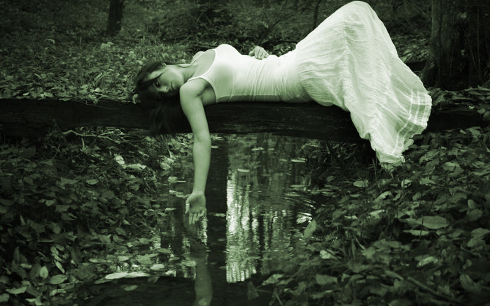A gothic model in a flowing white dress lies on a log above a tranquil water surface, surrounded by dark foliage, evoking a serene yet mysterious atmosphere.