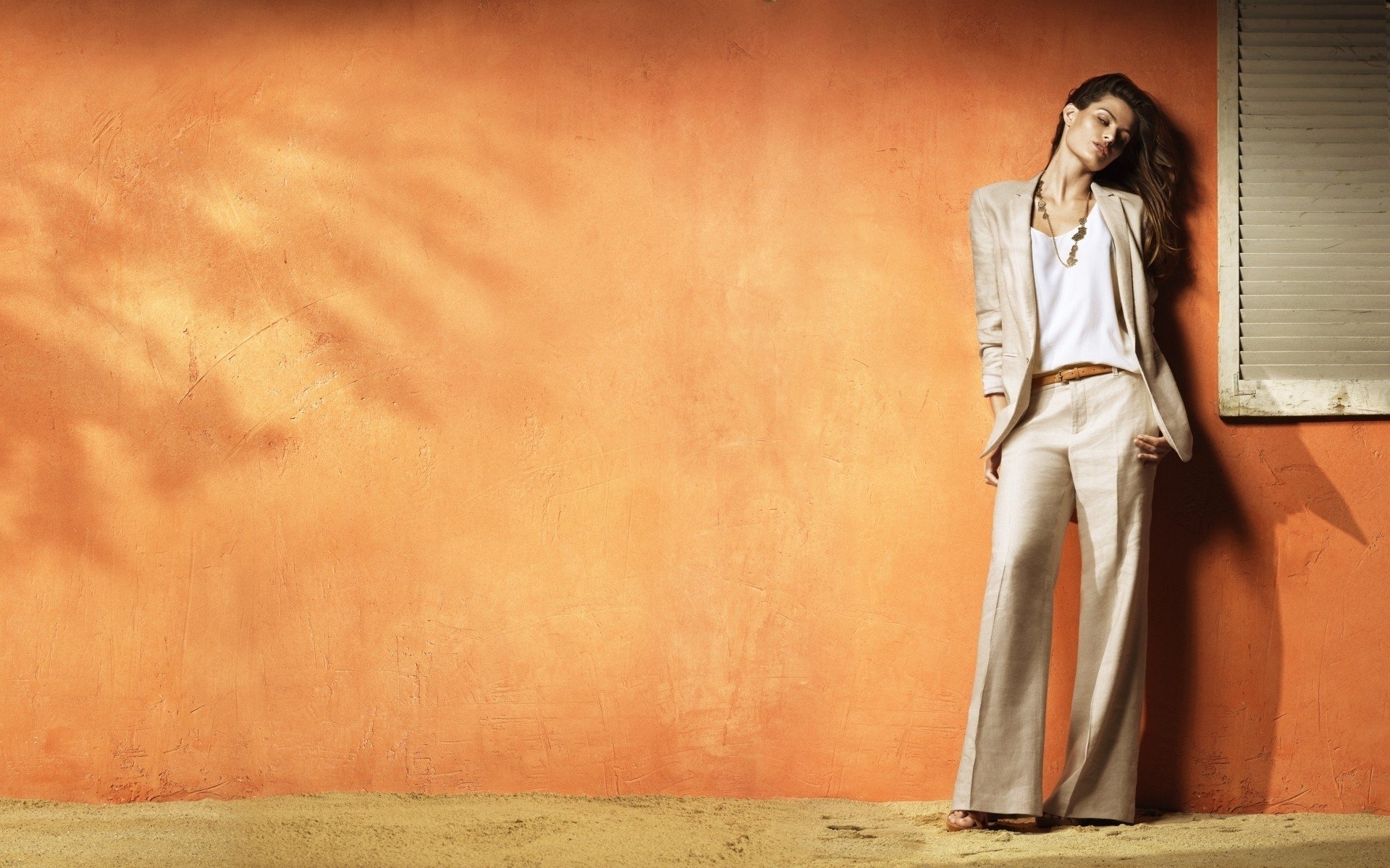 HD PC desktop wallpaper featuring a woman model in a light suit leaning against an orange textured wall with a window to the side.
