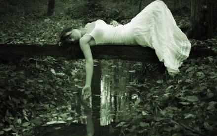A gothic model in a flowing white dress lies on a log above a tranquil water surface, surrounded by dark foliage, evoking a serene yet mysterious atmosphere.