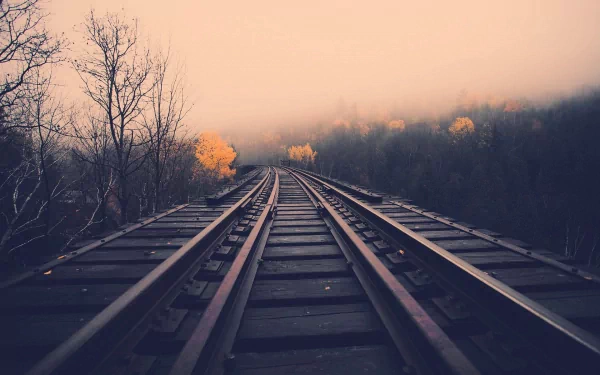 A serene HD desktop wallpaper featuring a winding railroad track disappearing into a foggy landscape, accented by autumn foliage and bare trees.