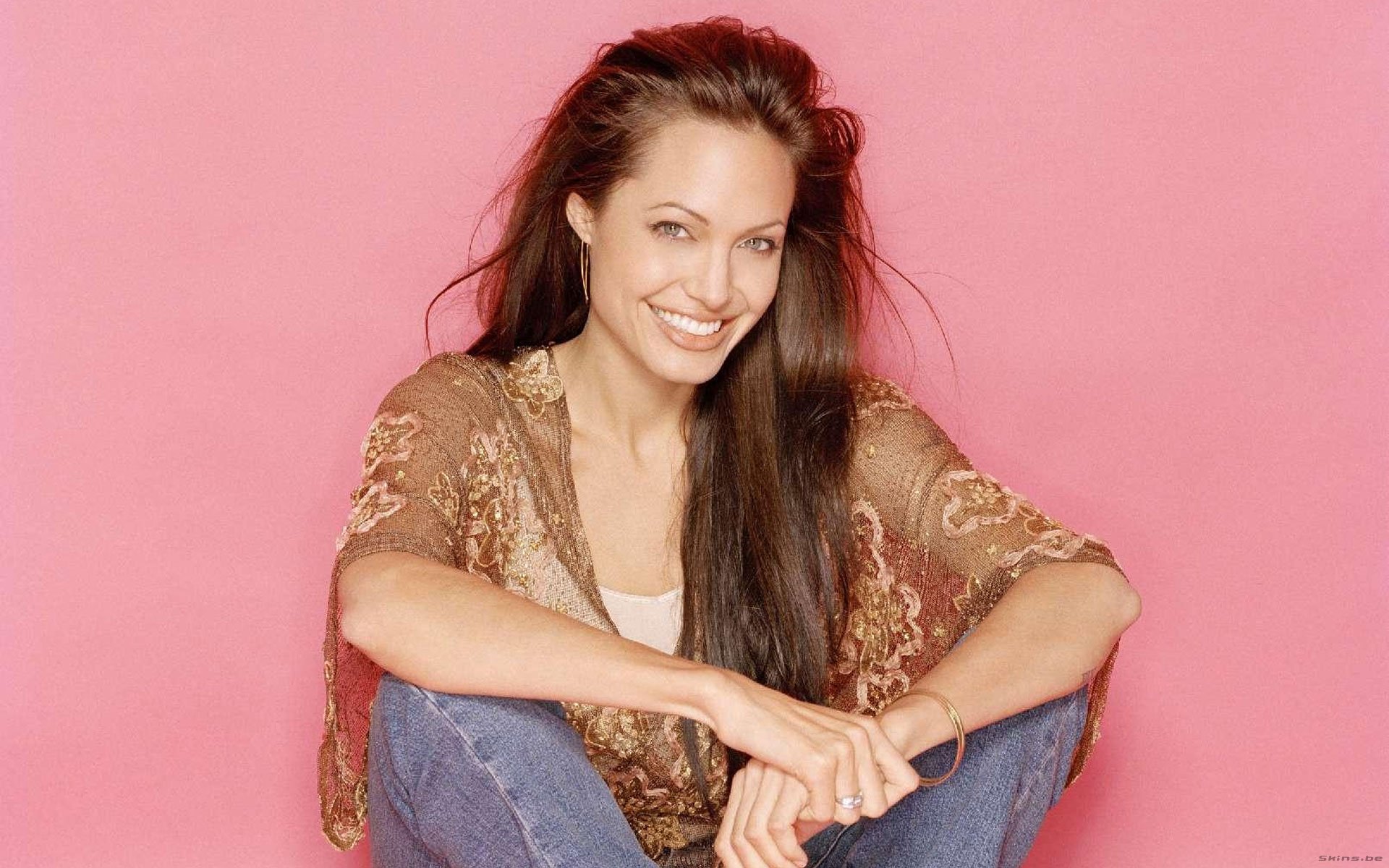 Angelina Jolie smiles warmly while sitting against a bright pink background, showcasing her effortless charm in this HD desktop wallpaper.