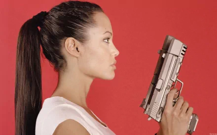 A striking HD wallpaper featuring Angelina Jolie in profile, holding a gun against a vibrant red background, capturing her fierce and commanding presence.