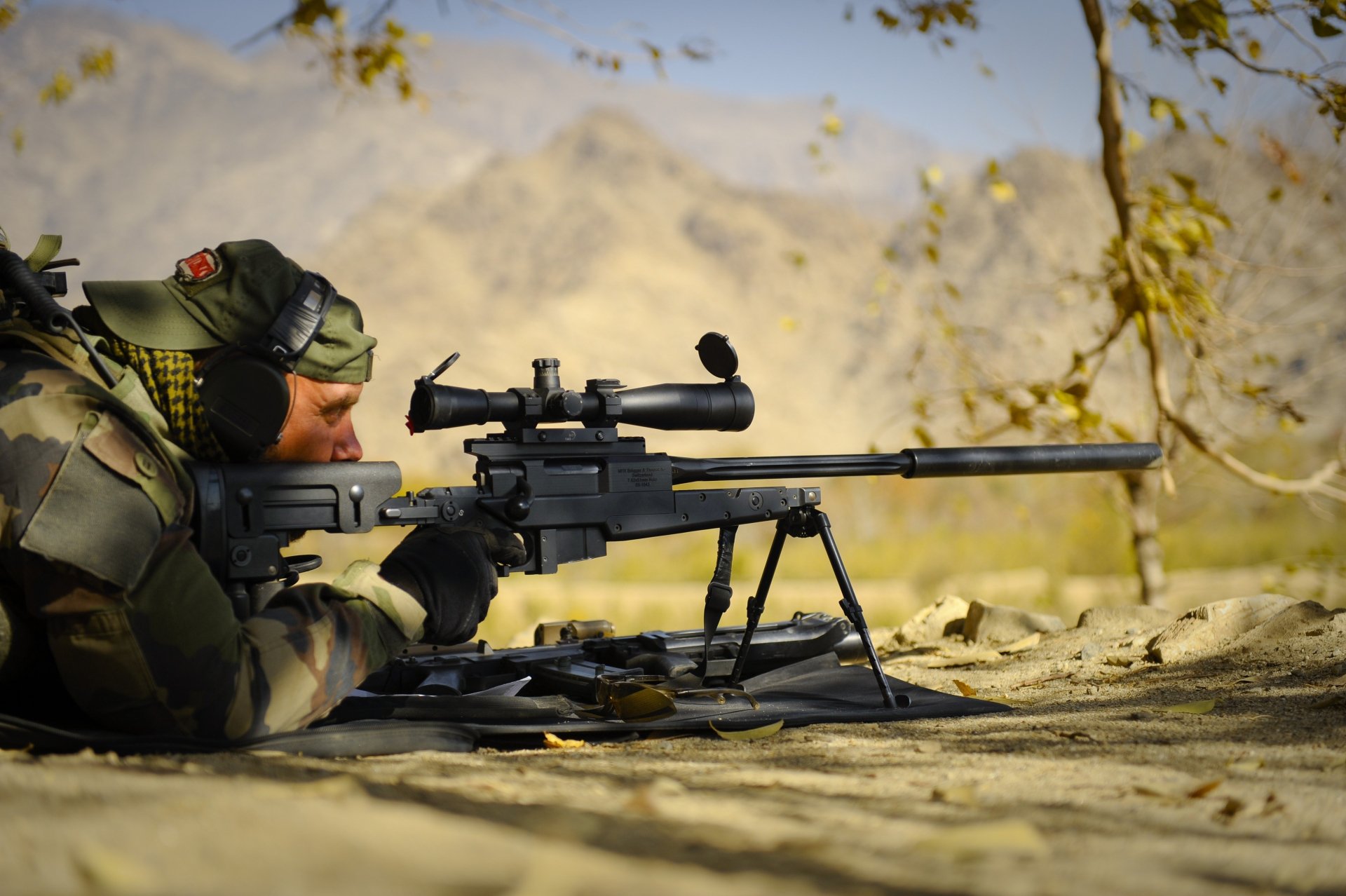 A military sniper in camouflage gear aims through a high-powered rifle on a bipod, set against a mountainous landscape in this 4K Ultra HD PC desktop wallpaper.