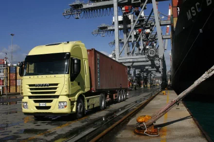 HD desktop wallpaper featuring a yellow Iveco vehicle transporting a shipping container at a busy industrial port.