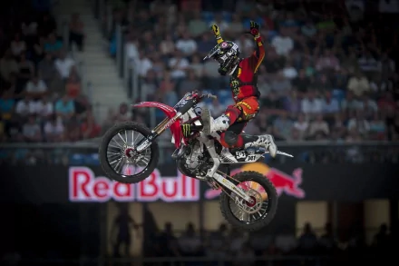 A motocross rider performs a daring aerial stunt in front of a crowd, captured in a high-definition desktop wallpaper. The vibrant scene includes a prominent Red Bull logo in the background.
