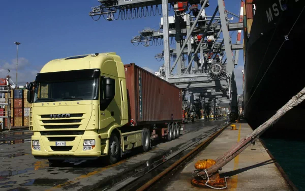 HD desktop wallpaper featuring a yellow Iveco vehicle transporting a shipping container at a busy industrial port.