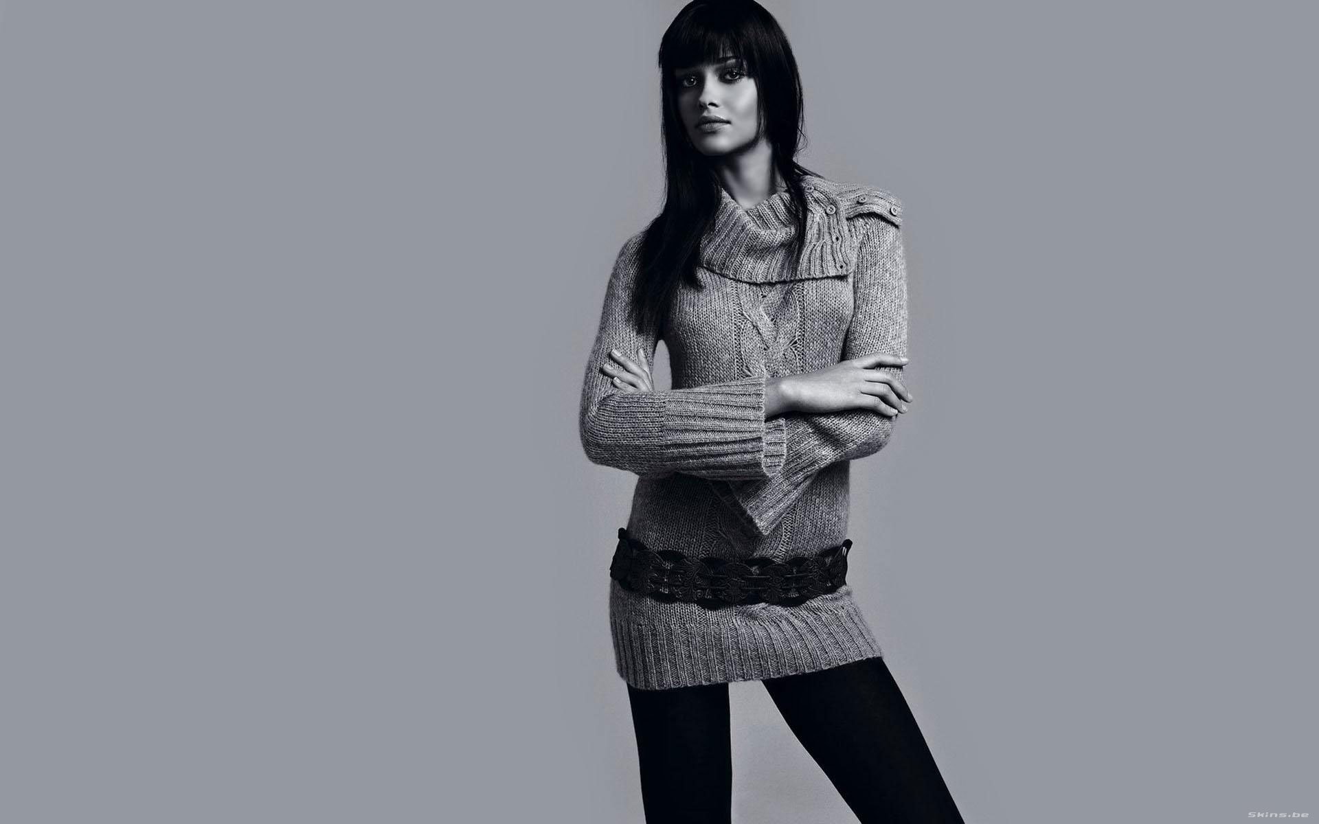 HD PC desktop wallpaper featuring a woman with long dark hair wearing a knit sweater dress and belt, standing against a plain gray background.