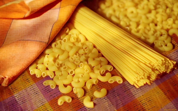 HD desktop wallpaper showing uncooked elbow macaroni and spaghetti pasta on a colorful cloth with an orange and cream checkered napkin nearby.