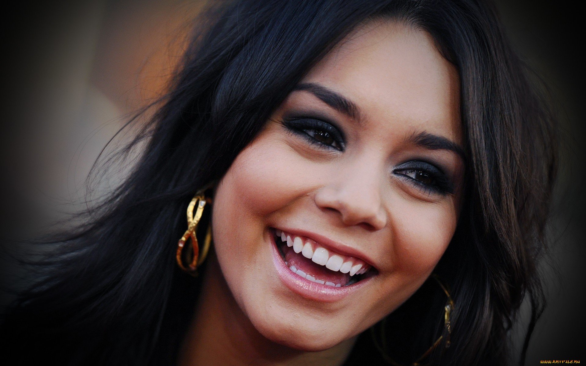 Close-up HD desktop wallpaper featuring celebrity Vanessa Hudgens smiling, with dark hair and gold hoop earrings against a blurred background.