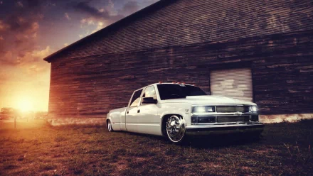 HD desktop wallpaper featuring a sleek, lowered white Chevy truck parked beside a rustic wooden barn at sunset.