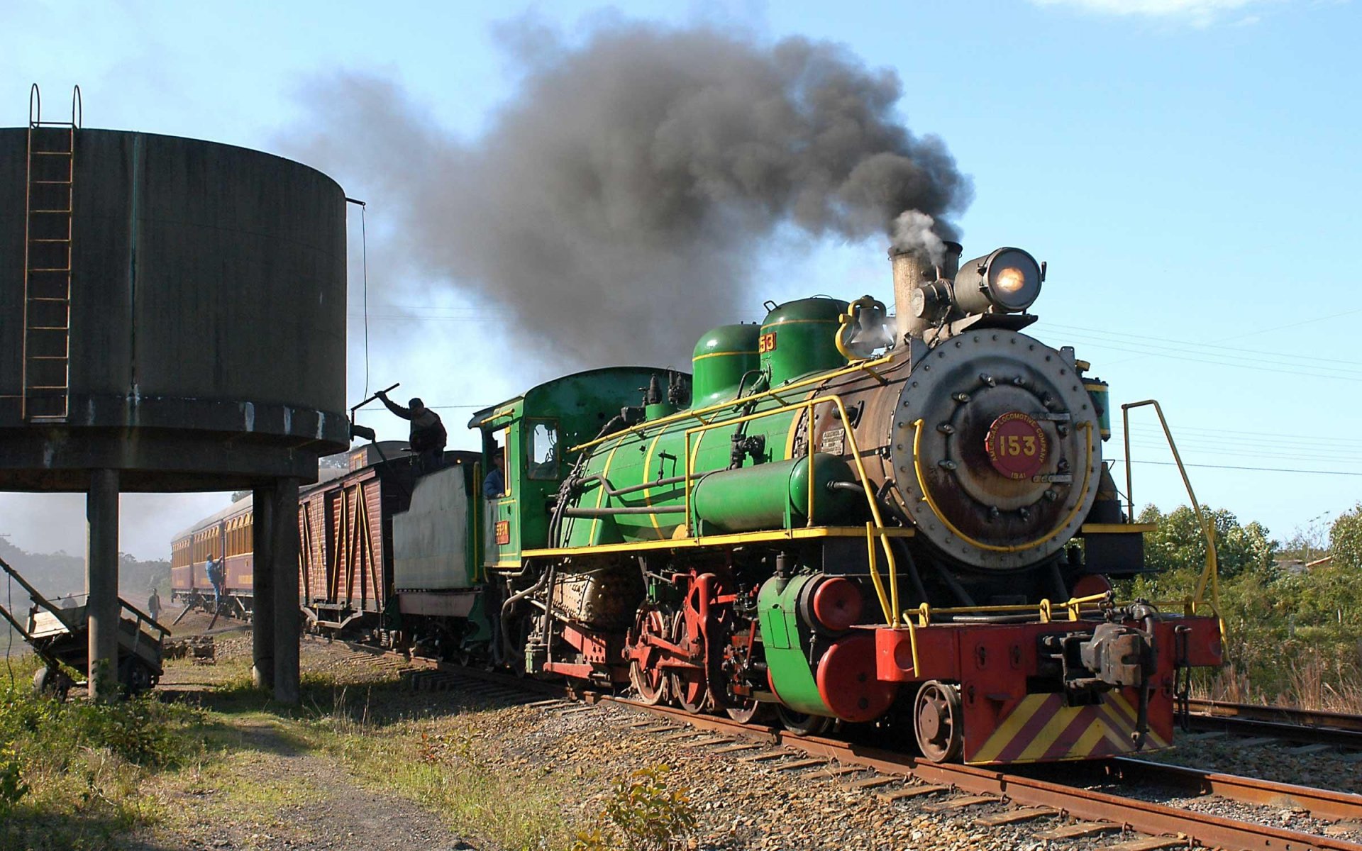 HD PC desktop wallpaper background: vintage green steam train (vehicle) billowing dark smoke as it rolls past a water tower on sunlit tracks.
