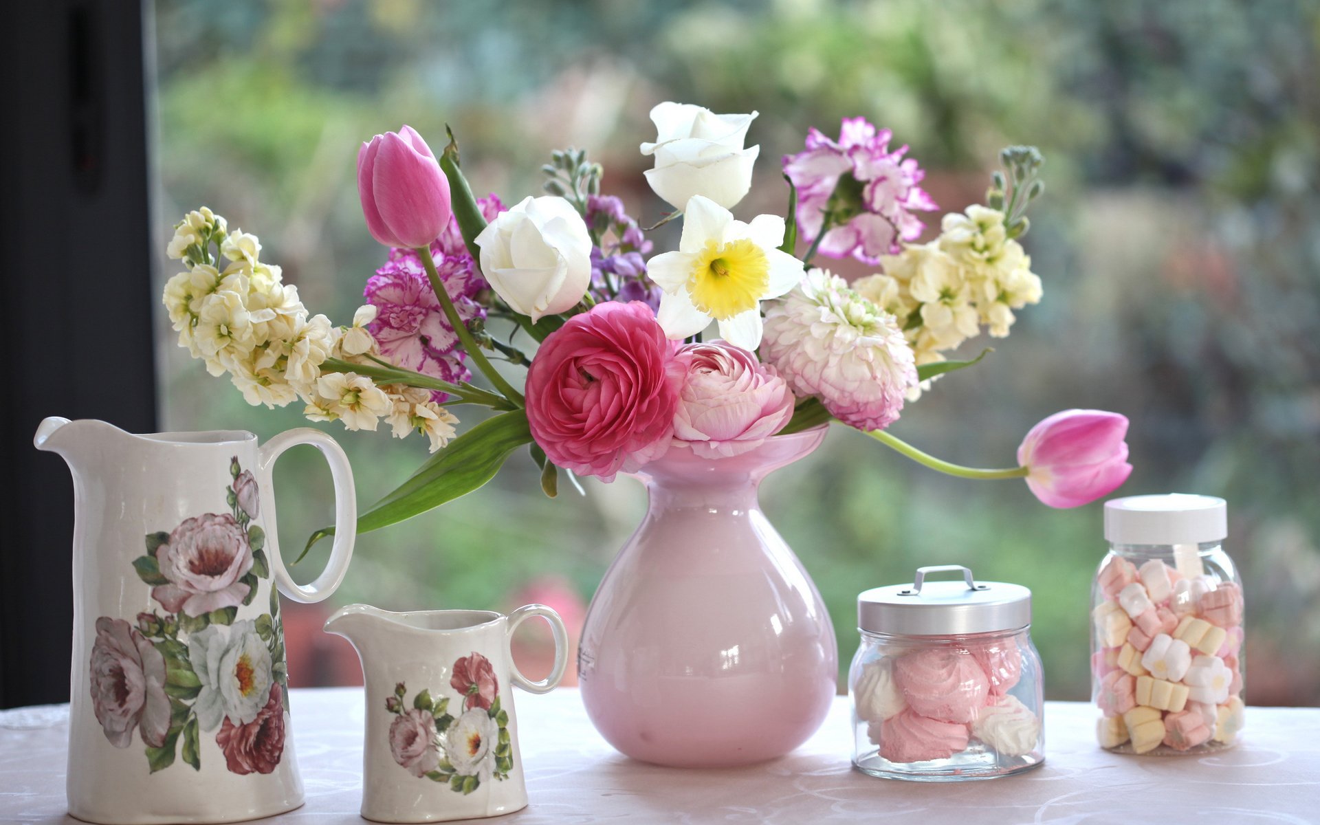 A still life HD desktop wallpaper featuring a bouquet of tulip, daffodil, ranuncula, carnation, and marshmallow flowers arranged in a pink pitcher vase on a windowsill.