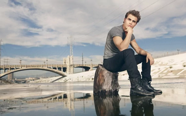 HD PC desktop wallpaper featuring celebrity Paul Wesley sitting thoughtfully on a rock by a reflective water surface with a bridge and cloudy sky in the background.