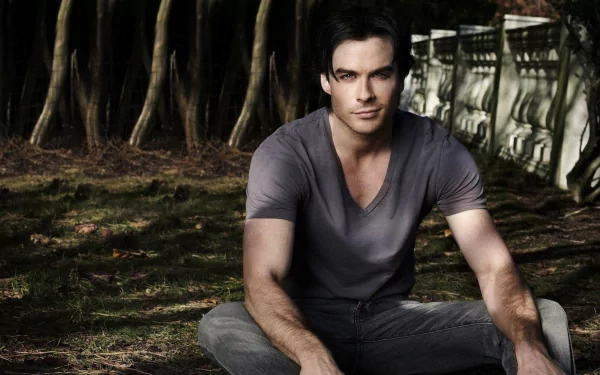 A striking HD wallpaper featuring Ian Somerhalder sitting in a natural setting, exuding a relaxed yet captivating presence against a backdrop of trees and greenery.