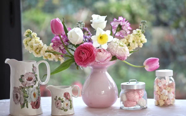 A still life HD desktop wallpaper featuring a bouquet of tulip, daffodil, ranuncula, carnation, and marshmallow flowers arranged in a pink pitcher vase on a windowsill.