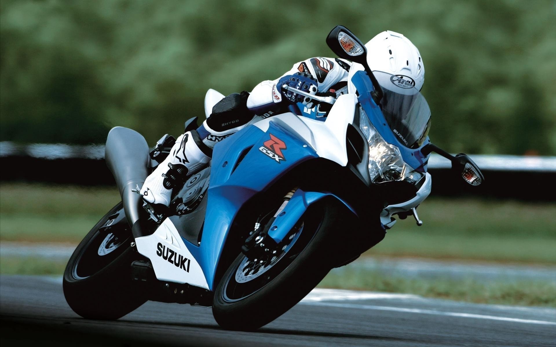 HD PC desktop wallpaper of a blue Suzuki sport motorcycle vehicle leaning sharply through a racetrack turn, rider in white leathers and helmet, blurred green background.