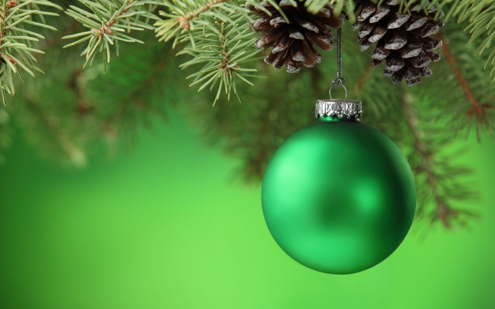 A vibrant green Christmas ornament hangs from pine branches, accompanied by pine cones, set against a soft green background, creating a festive holiday atmosphere.