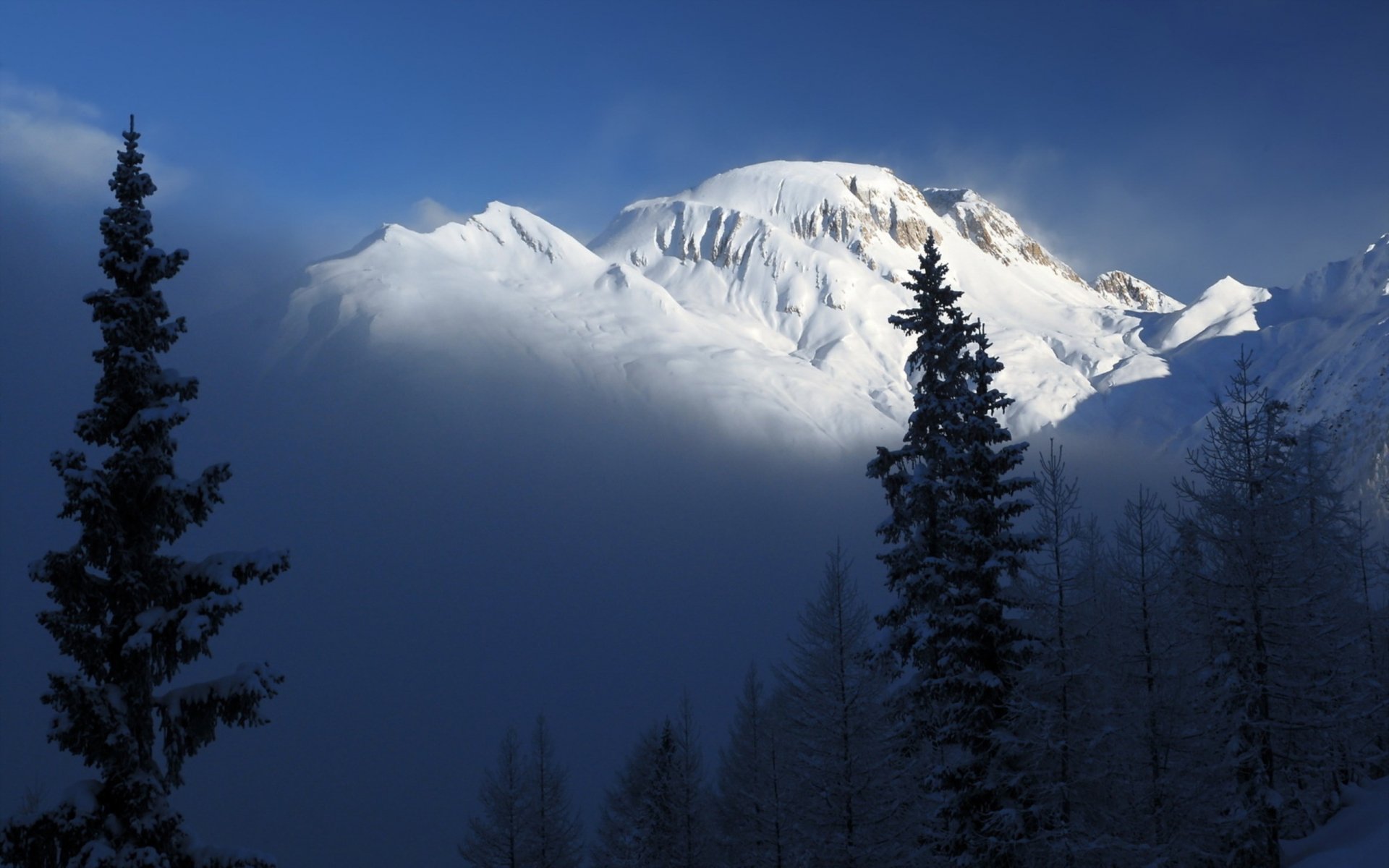 A stunning mountain landscape shrouded in mist, featuring snow-covered peaks and tall evergreen trees, captured in high definition for a captivating desktop wallpaper.