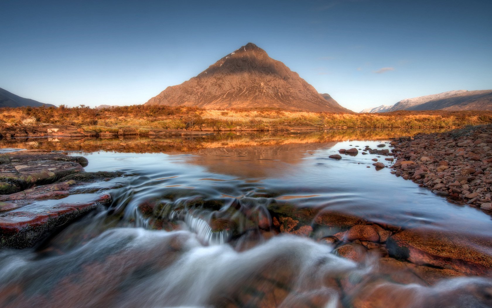 HD PC desktop wallpaper: sunrise-lit conical mountain reflected in a calm pool, foreground stream cascading over rocks under a clear blue sky.