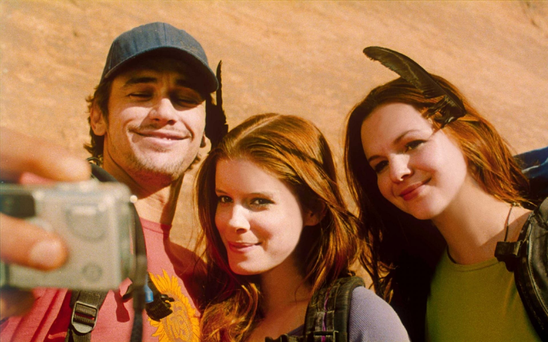 Three smiling people take a desert selfie against red rock cliffs, warm cinematic tones in a 2K Quad HD movie-style desktop wallpaper.