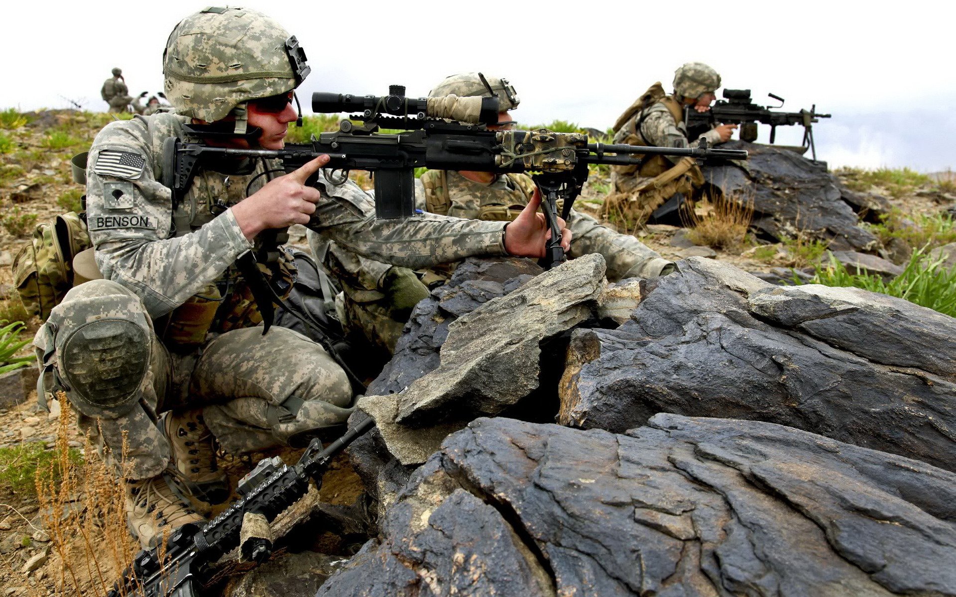 HD PC desktop wallpaper showing military soldiers in camouflage uniforms aiming rifles while positioned behind rocky terrain.
