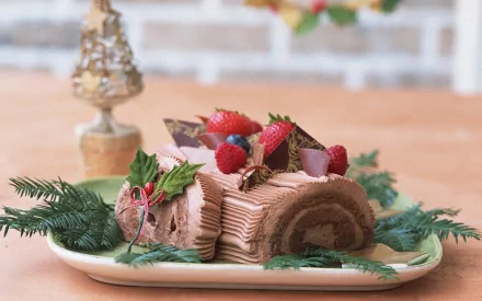 A beautifully presented chocolate Yule log dessert adorned with fresh raspberries and decorative chocolate, surrounded by greenery, making a delightful food-themed HD desktop wallpaper.