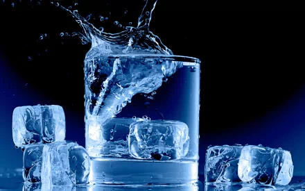 A high-definition photograph featuring an ice cube splashing into a glass of water, surrounded by additional ice cubes, set against a dark, blurred background.