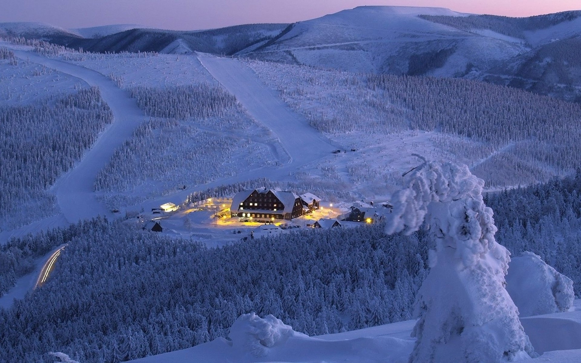 Winter photography HD PC desktop wallpaper: snow-clad mountains and forests at dusk, a warmly lit lodge in a snowy valley, foreground snow-burdened tree under a violet sky.