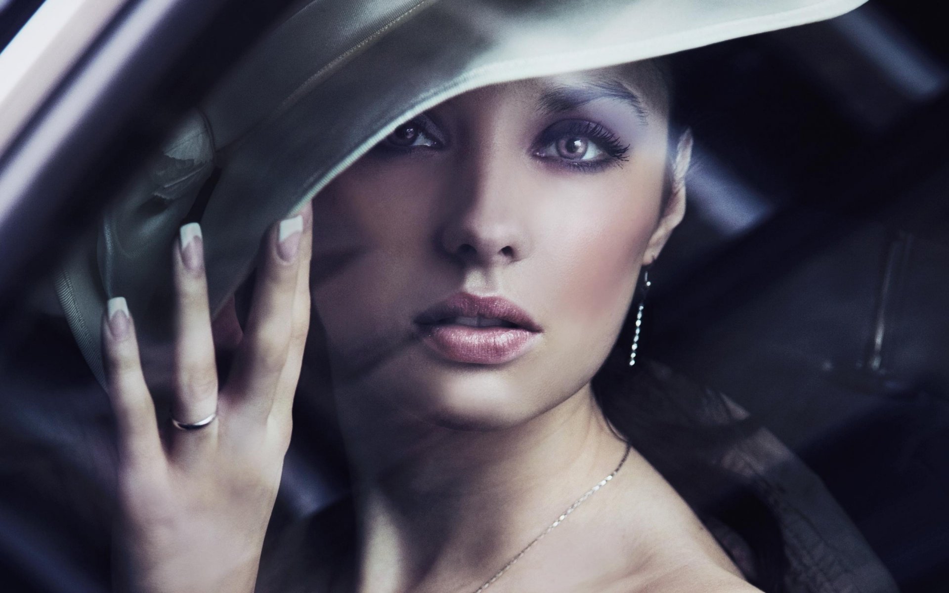 A captivating makeup model gazes through a car window, elegantly framed by her hat, showcasing her striking features and artistry in this high-definition wallpaper.