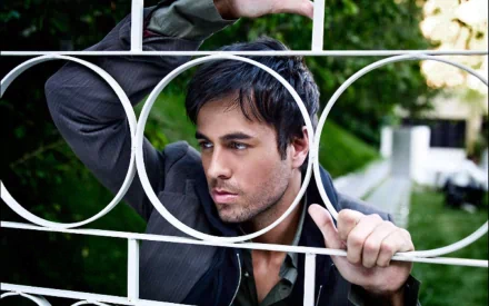 HD desktop wallpaper featuring Enrique Iglesias, captured thoughtfully with a contemplative expression as he leans against a decorative fence amid greenery.