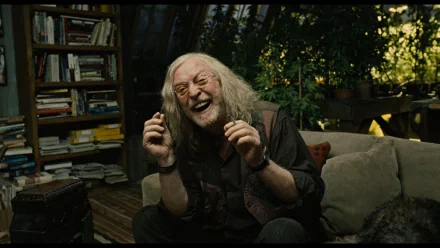 HD wallpaper featuring Michael Caine from Children Of Men, laughing in a cozy room filled with plants and books.