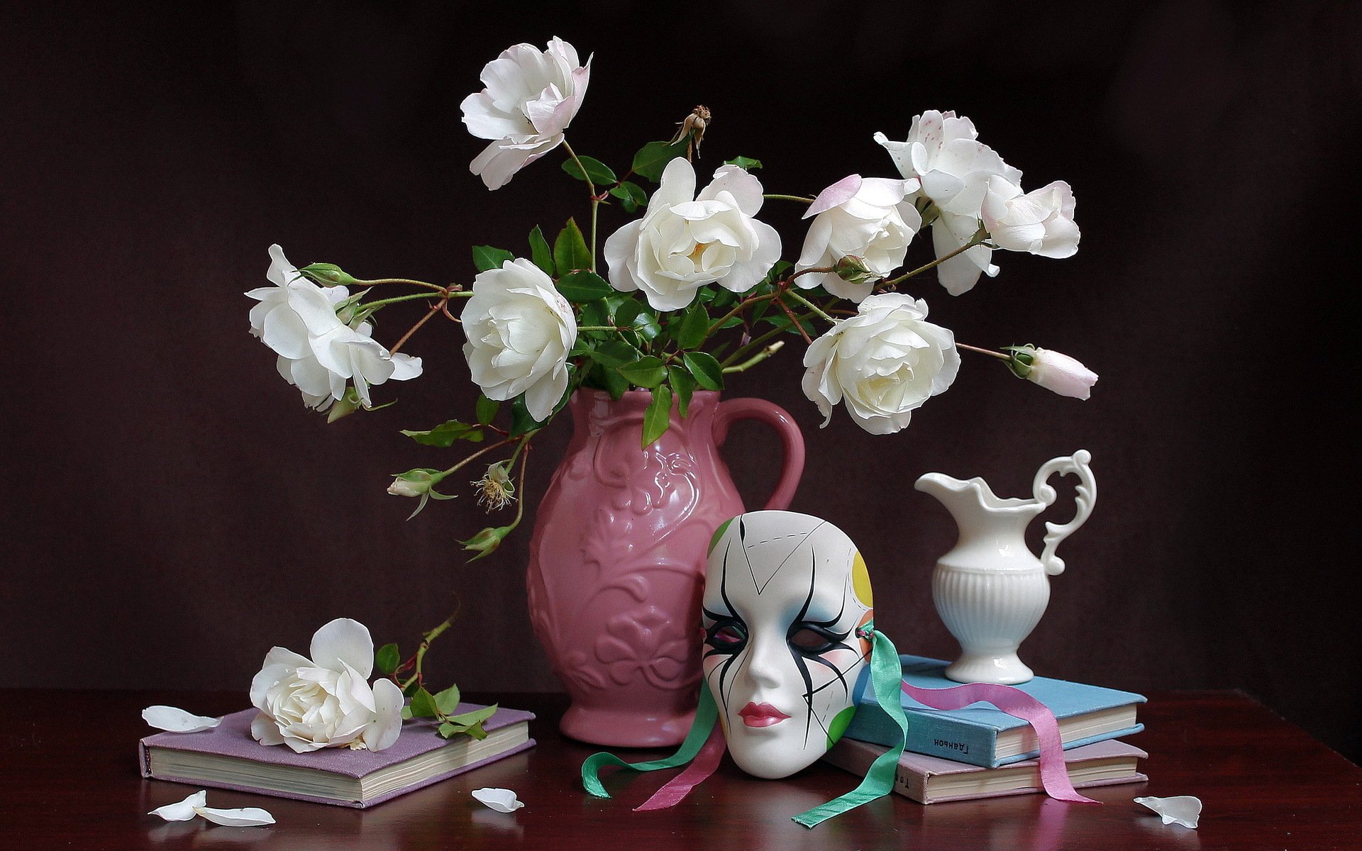 A serene still life featuring a pink vase with white roses, a decorative mask, and stacked books, set against a dark background, creating an elegant HD wallpaper.