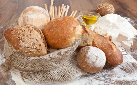 HD desktop wallpaper featuring various types of bread, a sack of flour, breadsticks, and olive oil on a wooden surface, highlighting baking and food themes.