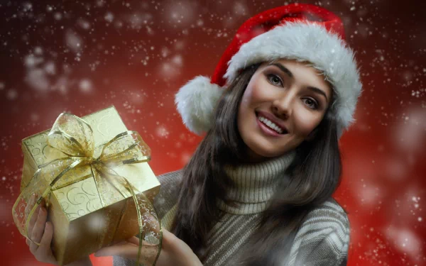 A smiling brunette woman in a Santa hat holds a wrapped gift against a festive red background, capturing the joyful spirit of the holiday season.