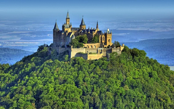 Hohenzollern Castle majestically sits atop a lush green hill, surrounded by expansive landscapes, making for a stunning HD desktop wallpaper and background.