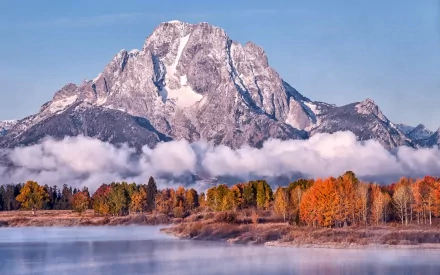 HD desktop wallpaper showcasing a majestic mountain rising above autumn trees with a layer of mist and clouds under a clear blue sky.