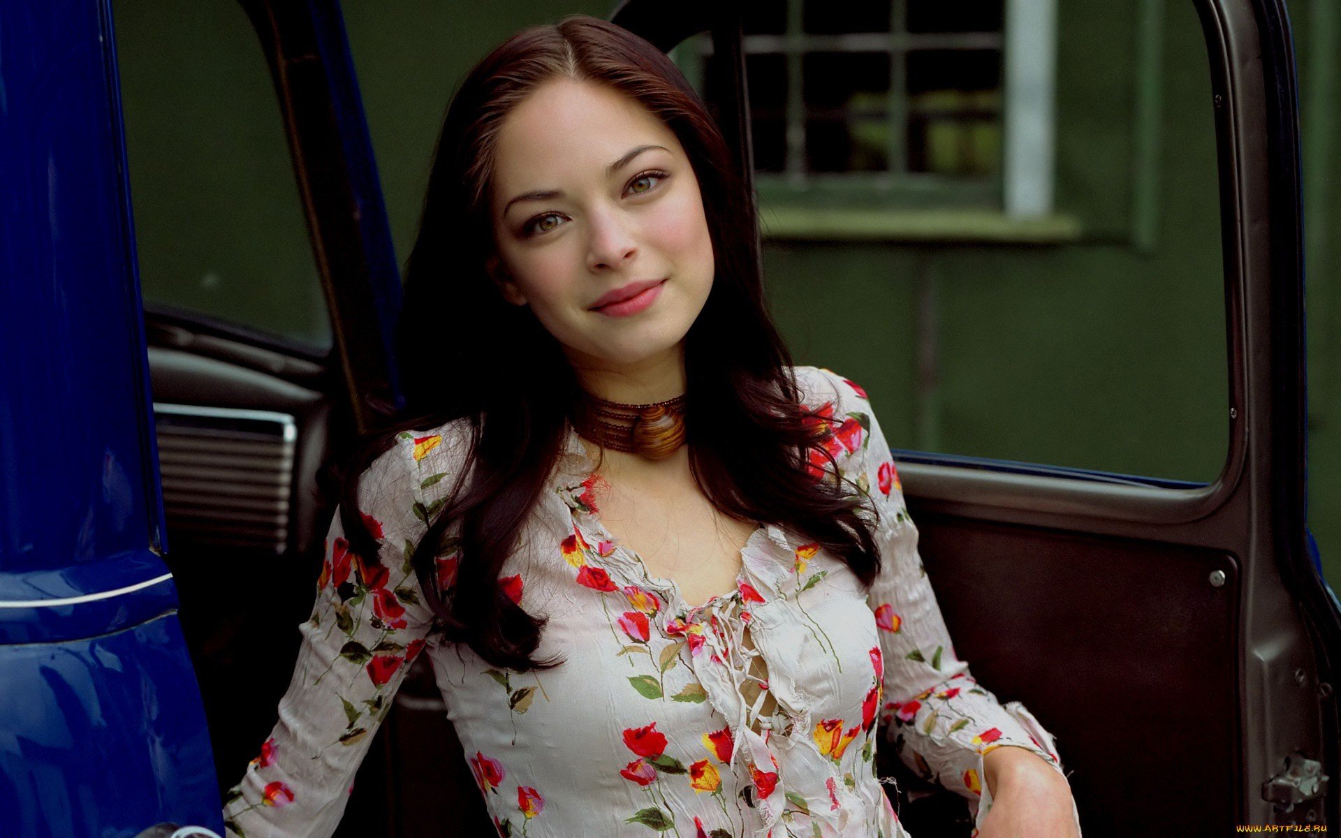 A captivating HD desktop wallpaper featuring Kristin Kreuk, showcasing her charming smile and floral-patterned dress against a rustic backdrop.