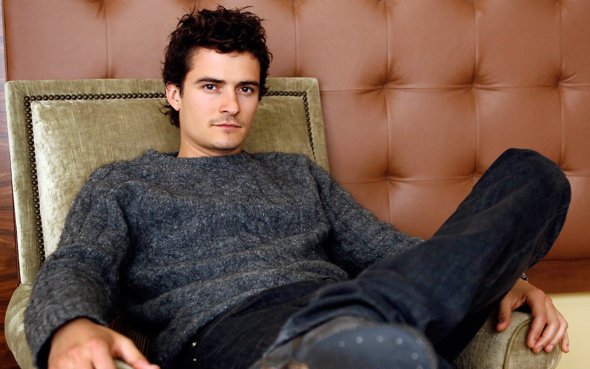 HD PC desktop wallpaper of a male celebrity reclining in an armchair, wearing a gray sweater and jeans against a tufted leather backdrop.