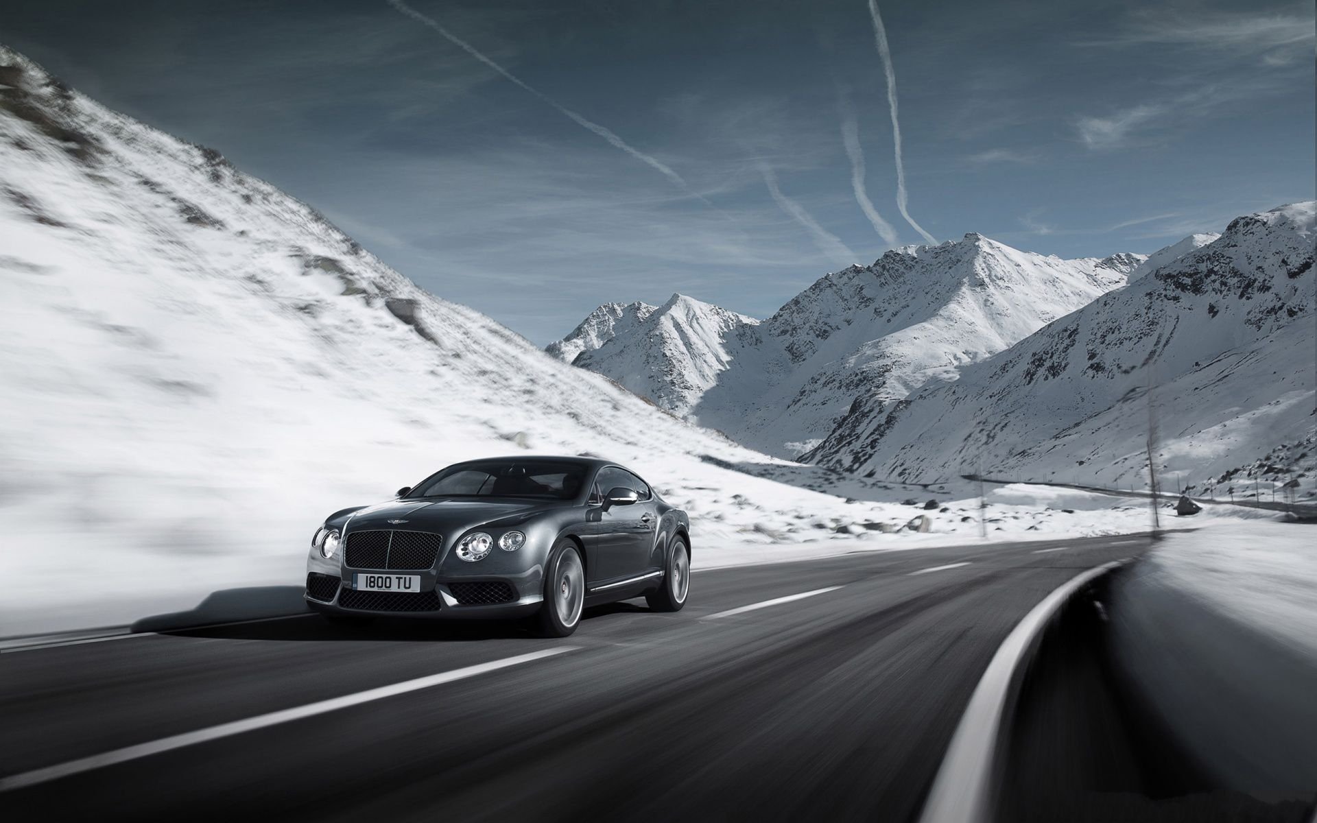 HD PC desktop wallpaper of a Bentley Continental vehicle cruising a winding mountain road, black Bentley against snowy alpine peaks and a dramatic sky.