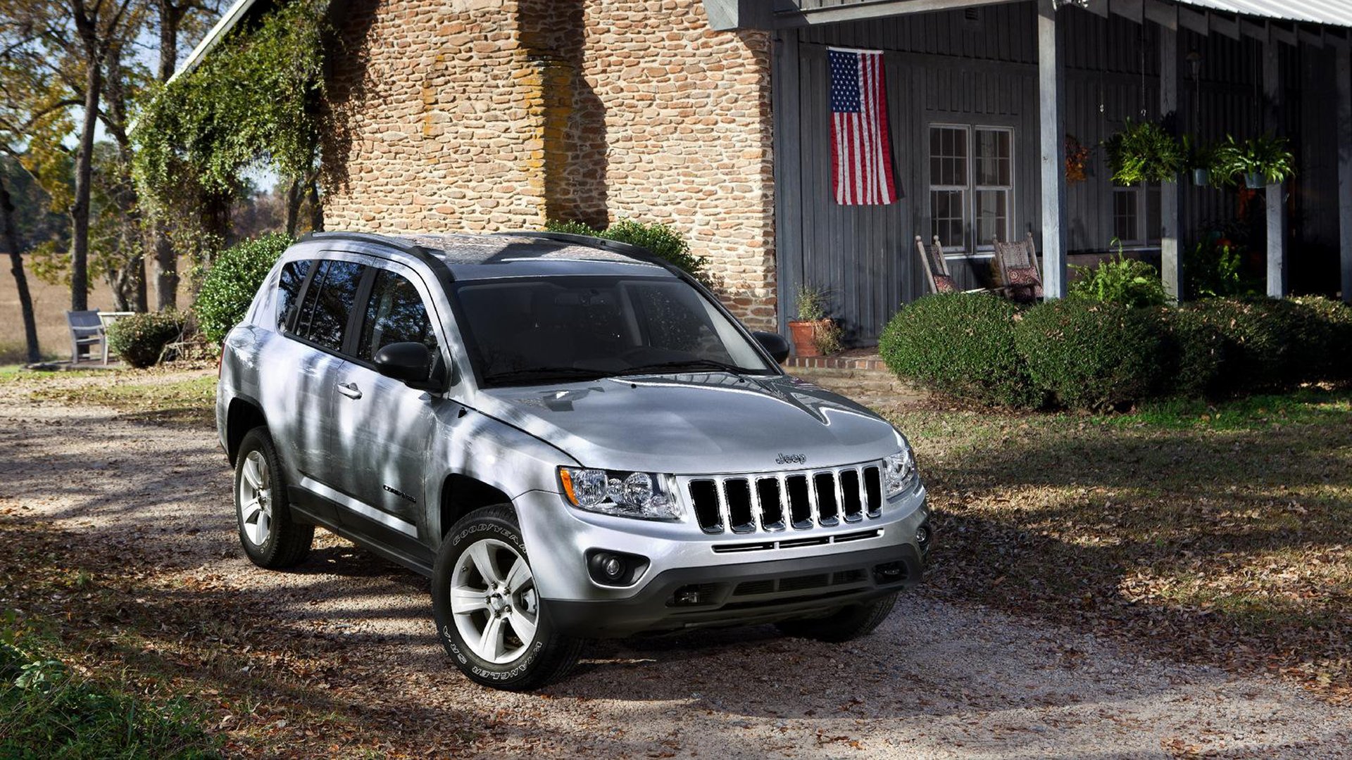A silver Jeep parked on a gravel driveway beside a stone house adorned with an American flag, surrounded by lush greenery, captures a tranquil outdoor scene. HD desktop wallpaper.