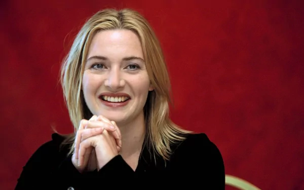 HD wallpaper featuring celebrity Kate Winslet sitting against a vibrant red background, smiling warmly with hands clasped.