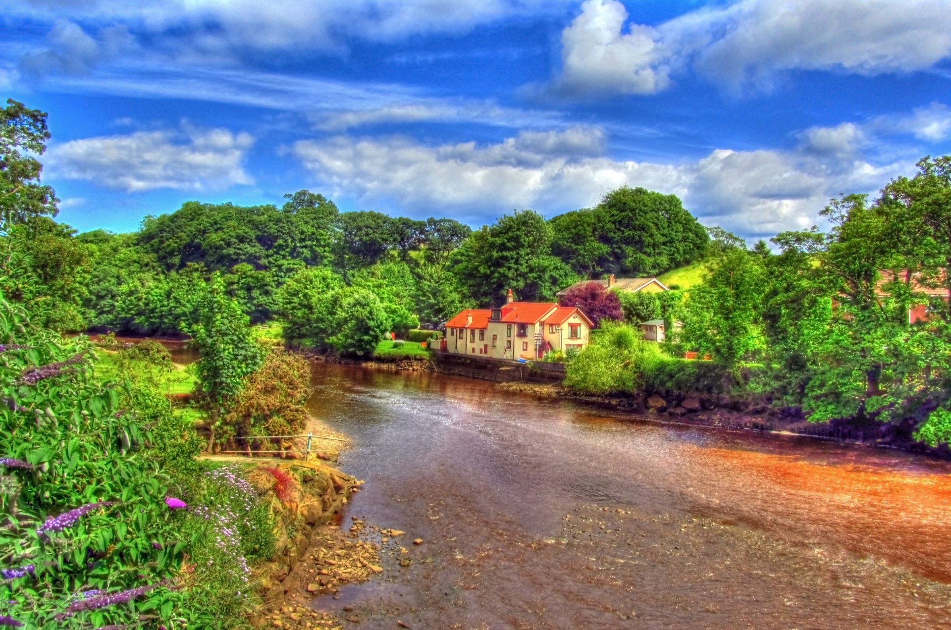 A serene river flows through lush greenery, with a charming house nestled against a backdrop of vibrant clouds and a clear sky, captured in stunning HDR photography.