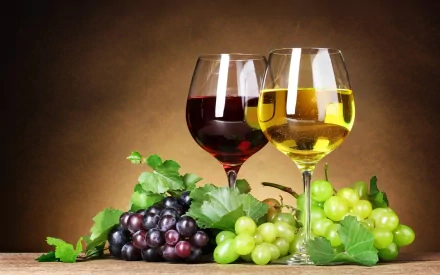 HD desktop wallpaper featuring two wine glasses—one with red wine and one with white—surrounded by fresh green and purple grapes and green leaves against a warm background.