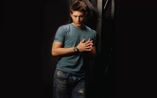 Jensen Ackles stands confidently in a casual blue shirt and jeans, hands clasped, set against a dark background, creating a striking HD desktop wallpaper.