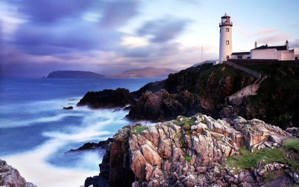 HD PC desktop wallpaper: Ireland coast with a man-made lighthouse on rocky cliffs, waves crashing, a distant island and a violet dusk sky.