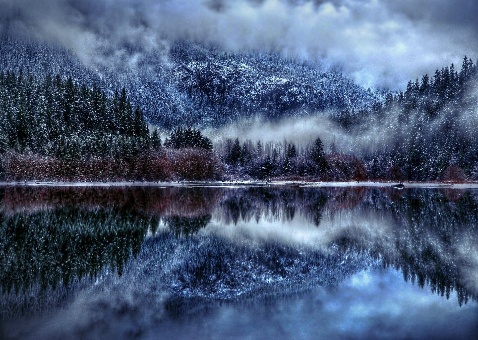 HD PC wallpaper featuring a serene nature scene with snow-covered trees and mountains perfectly reflected in a calm lake under a cloudy sky.