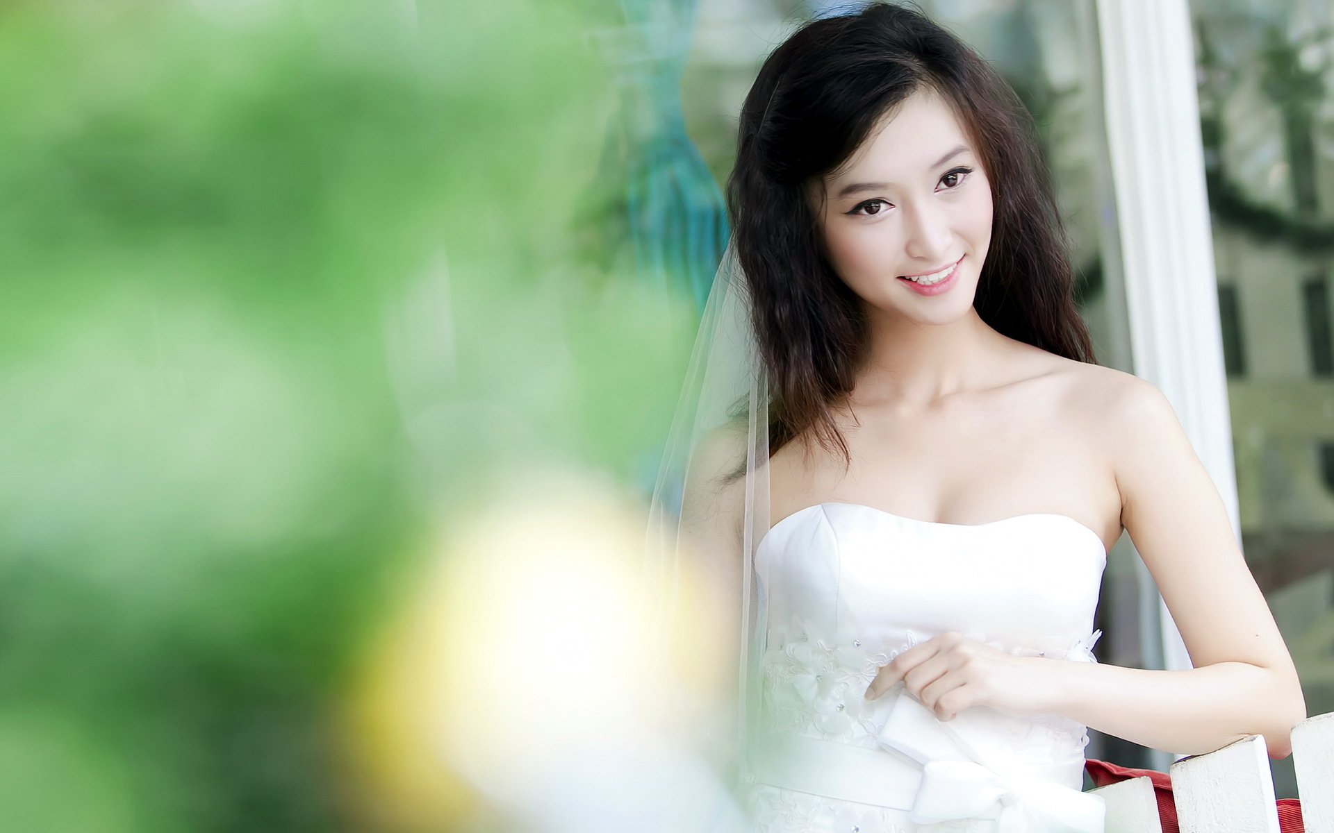 A smiling Asian woman in a white dress poses in a bright setting, surrounded by soft greenery. This HD wallpaper captures a serene and joyful moment.