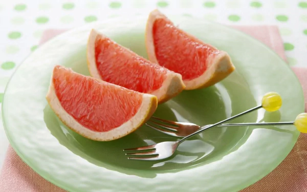 Three vibrant grapefruit wedges rest on a pale green plate, accompanied by two small forks, set against a soft polka dot background, creating a fresh and inviting desktop wallpaper.