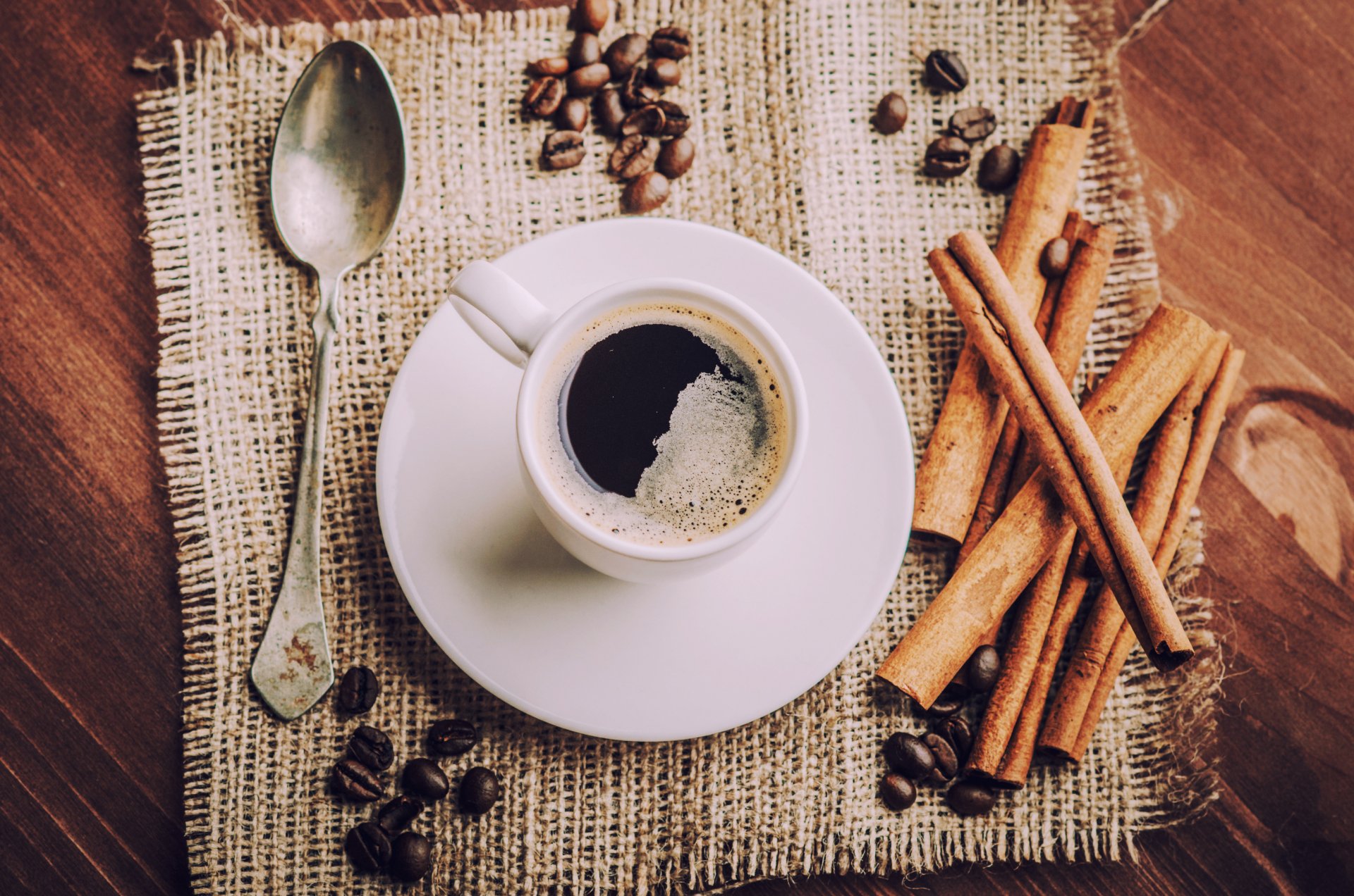 A cozy display of a coffee cup on a textured surface, surrounded by coffee beans, a spoon, and cinnamon sticks, creating a warm food-themed HD desktop wallpaper.
